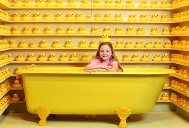 Rows of plastic ducks surrounding a yellow bath with a child in it and a duck on her head