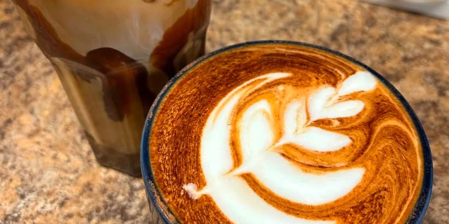 A coffee with a milk froth design on top and a glass of ice coffee served at Panama Café