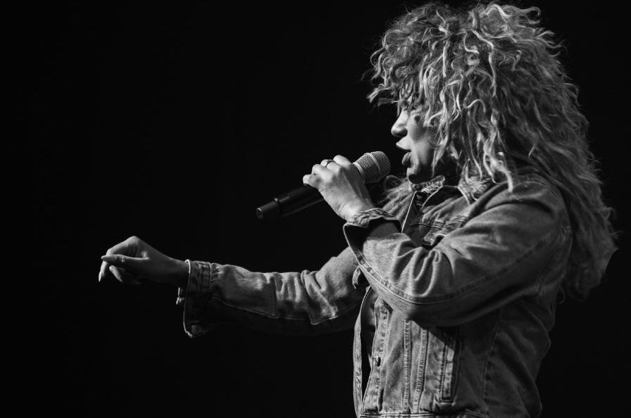 Side view of woman with big hair singing into a mic, against dark, plain background.