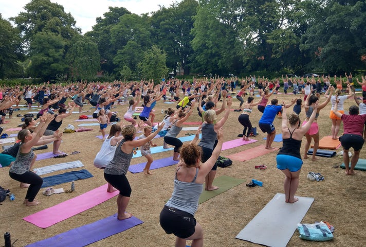 View of a large park area with hundreds of people spread out with yoga mats, all standing in a yoga pose.