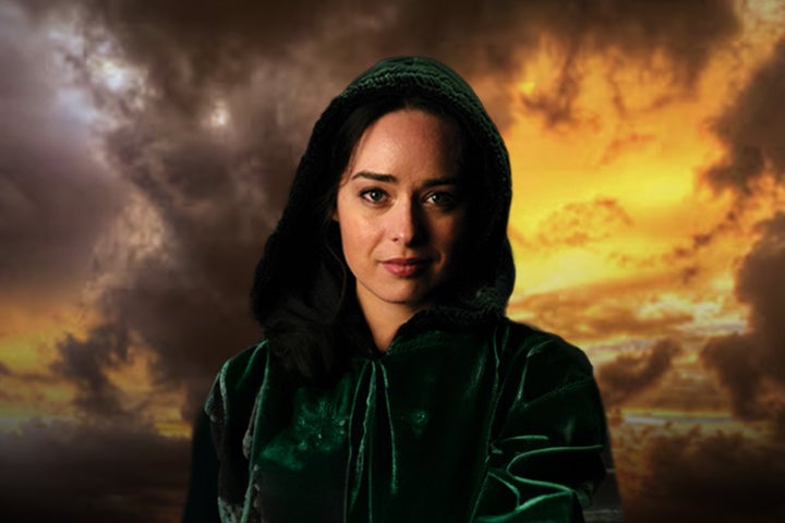 A woman is looking intently ahead in dark hooded cloak against dark, orange stormy sky.