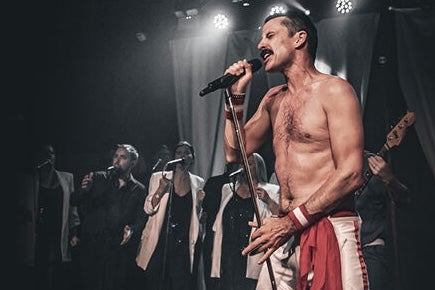 A man with no shirt on is singing with passion into a mic with singers in the background.