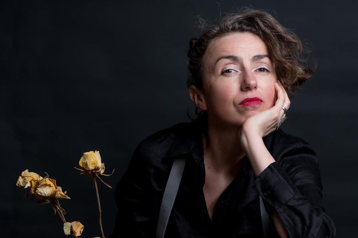 Against a black background, Clare, wearing black clothes, grey suspenders and red lipstick with her left hand under her chin, her left foot on a chair, is clasping a bottle and dead white roses with her right hand.