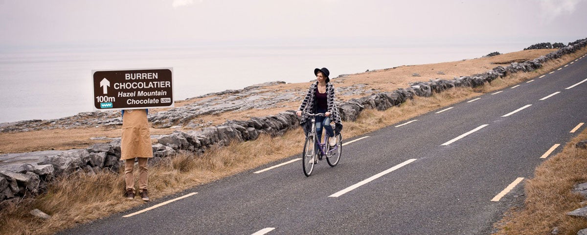 A woman cycling her bicycle along a coastal road while passing by a Burren Chocolatier road sign