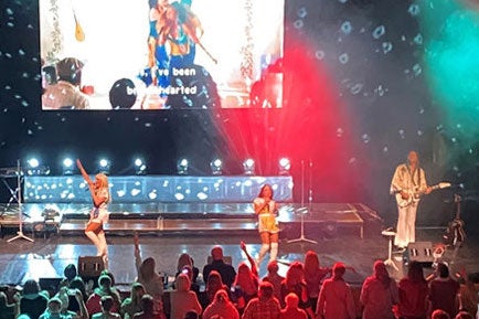 View of a stage looking over the audience with 2 women singing and 2 men playing instruments with large screen at the back of the stage.