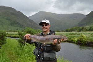 Man holding a fish, standing on the riverbank with mountains in the background