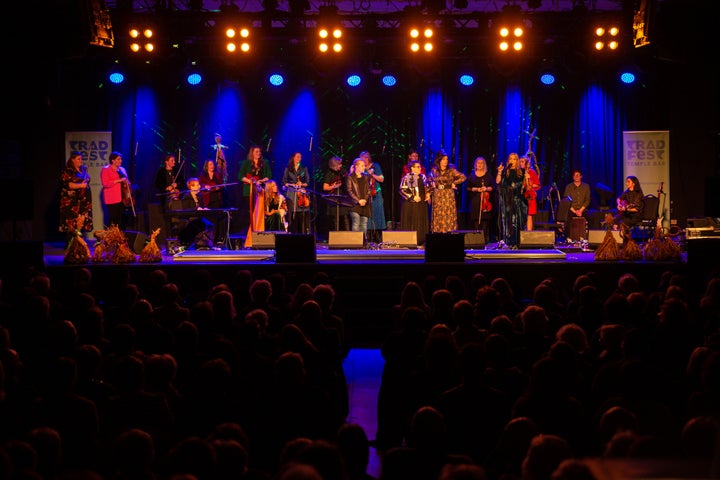 A stage seen from a distance with large group of women in 2 rows, singing and playing musical  instruments.