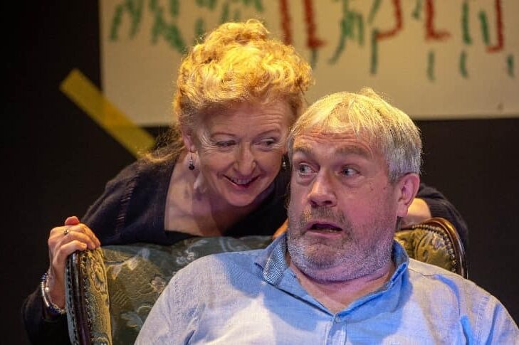 A woman is leaning over the back of an armchair smiling, saying something to a shocked looking man in blue shirt seated.