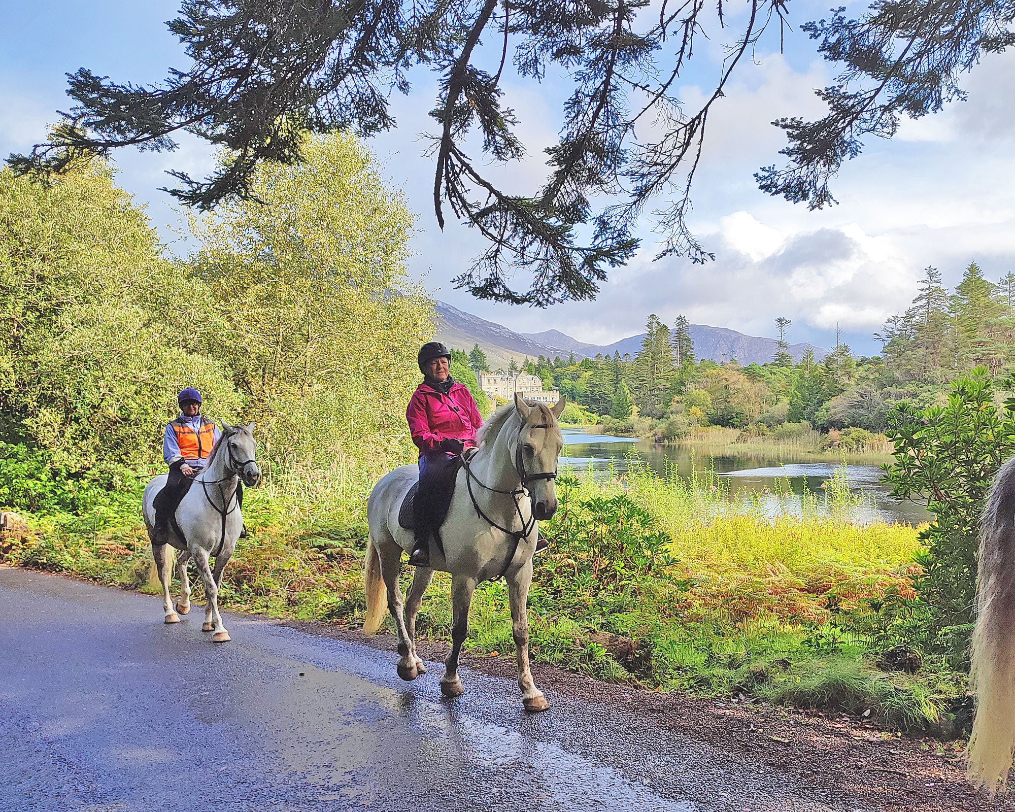 Connemara Equestrian Escapes view of two people on horseback with a lake and trees and mountains behind