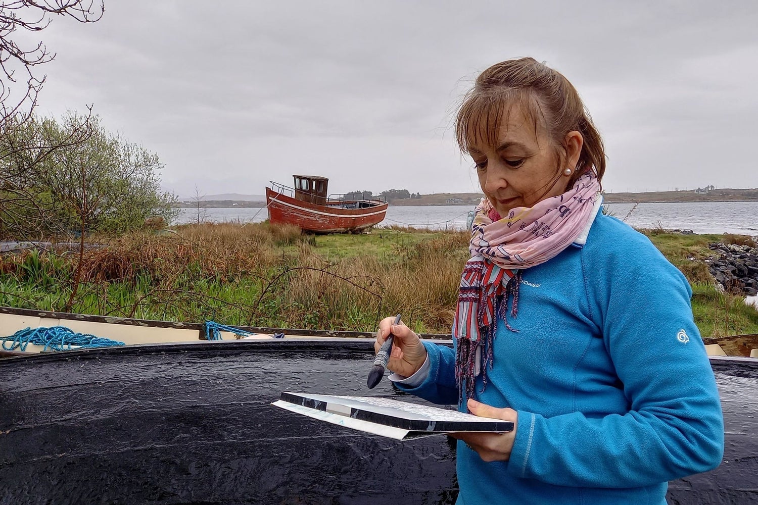 An artist with a pad and paintbrush in hand with an old boat in the background