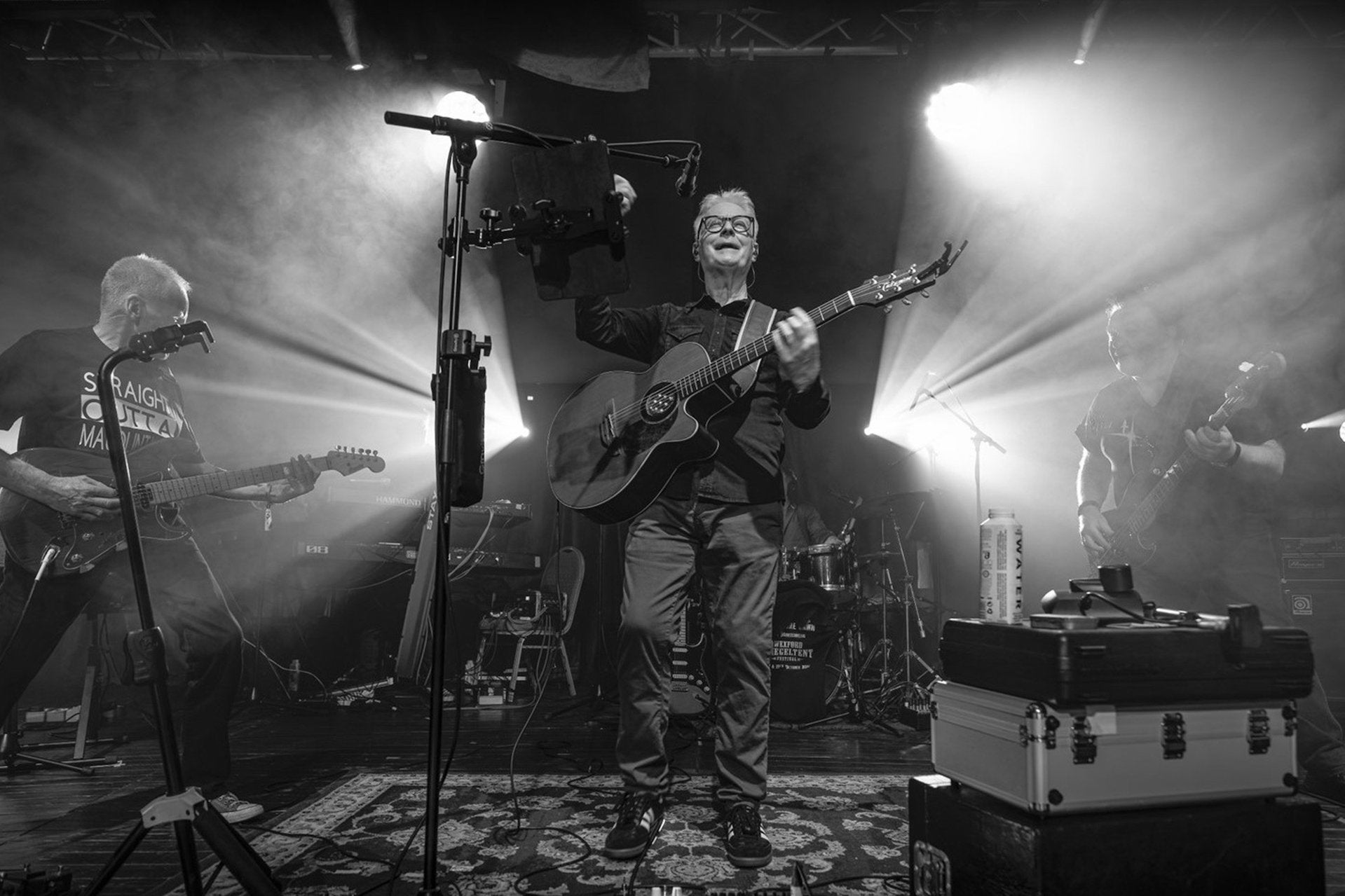 Black and white photo of man with guitar on a stage with 2 bright lights shining outwards either side of him.