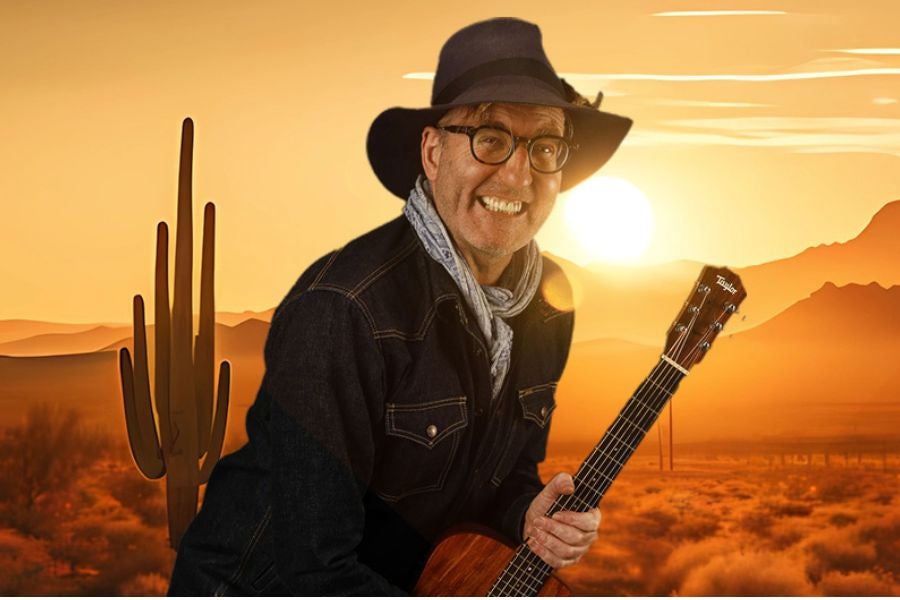 A grinning man in dark hat and jacket is clutching a guitar against backdrop of a desert with yellow and orange setting sun.