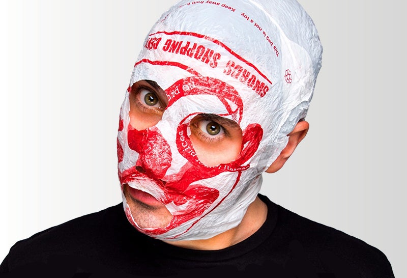 The face of a man is covered in a mask made from a white and red carrier bag, with eye and mouth holes cut away.