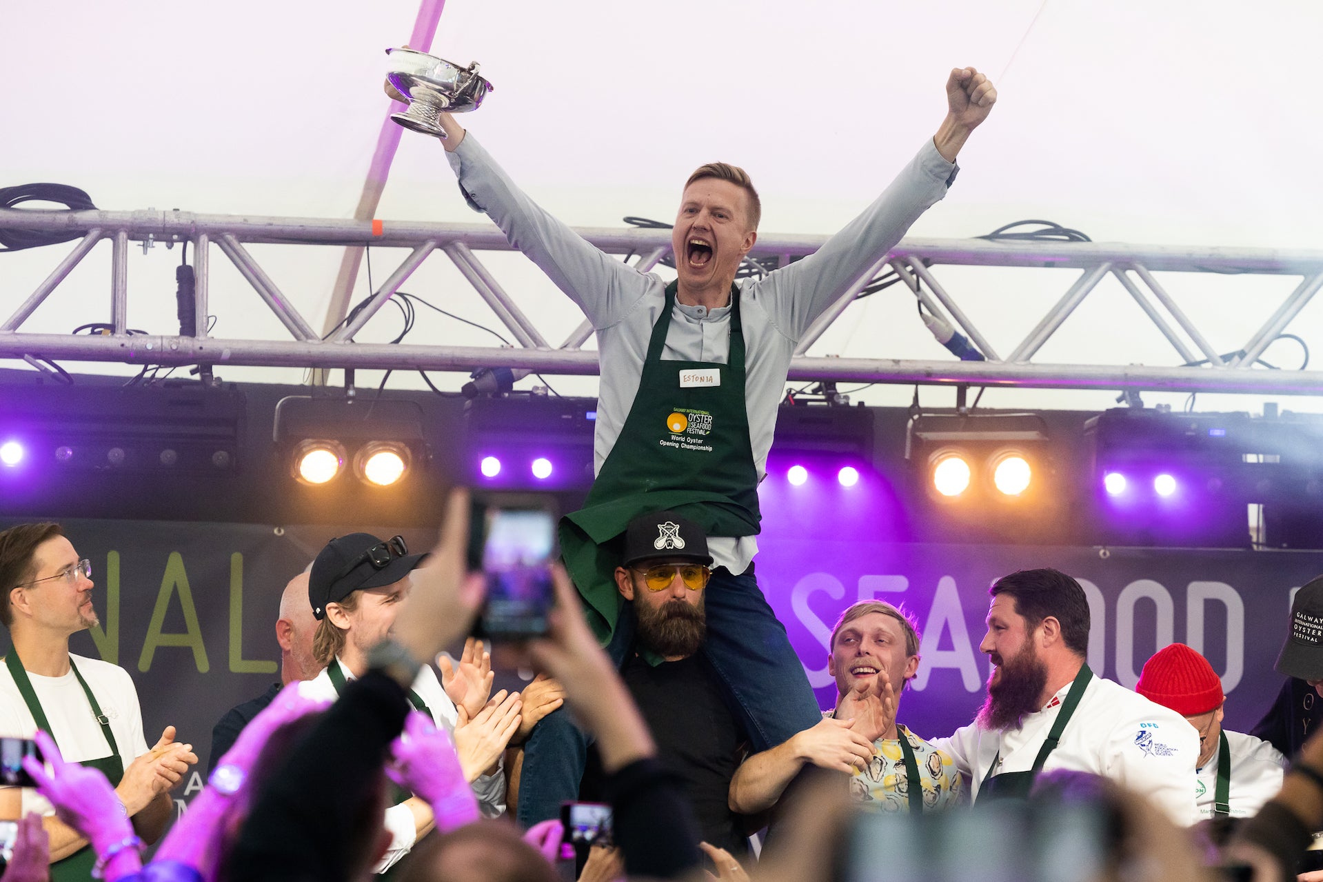 A man in black apron is celebrating with arms raised up, on the shoulders of other people with temporary event stage behind.