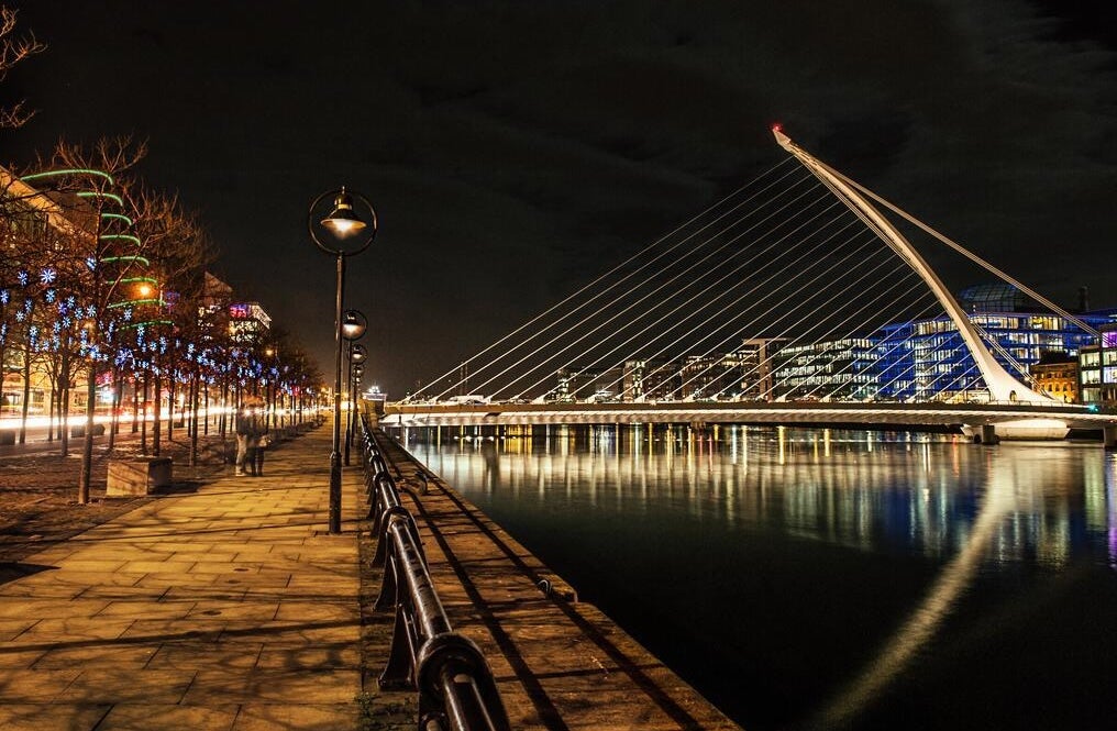 A view down alongside a river bank in the city at night with bridge lit up and buildings in the distance.