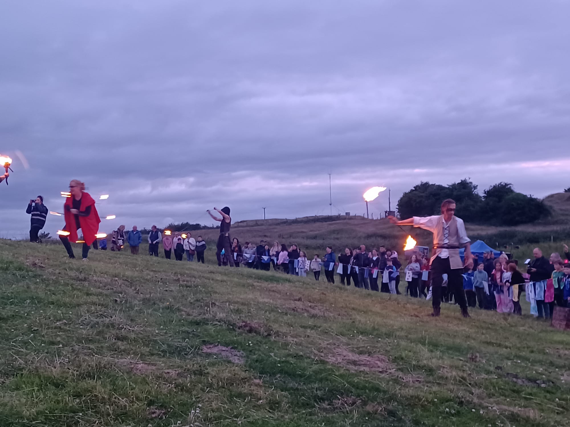 A large group of people in the distance on a hill at dusk, some holding lit flames.