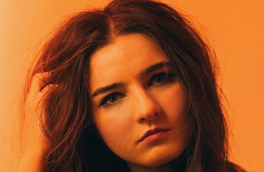 Brigit x District Muireann Bradley - headshot of Solomon woman with hand in her hair on on side, against orange background.