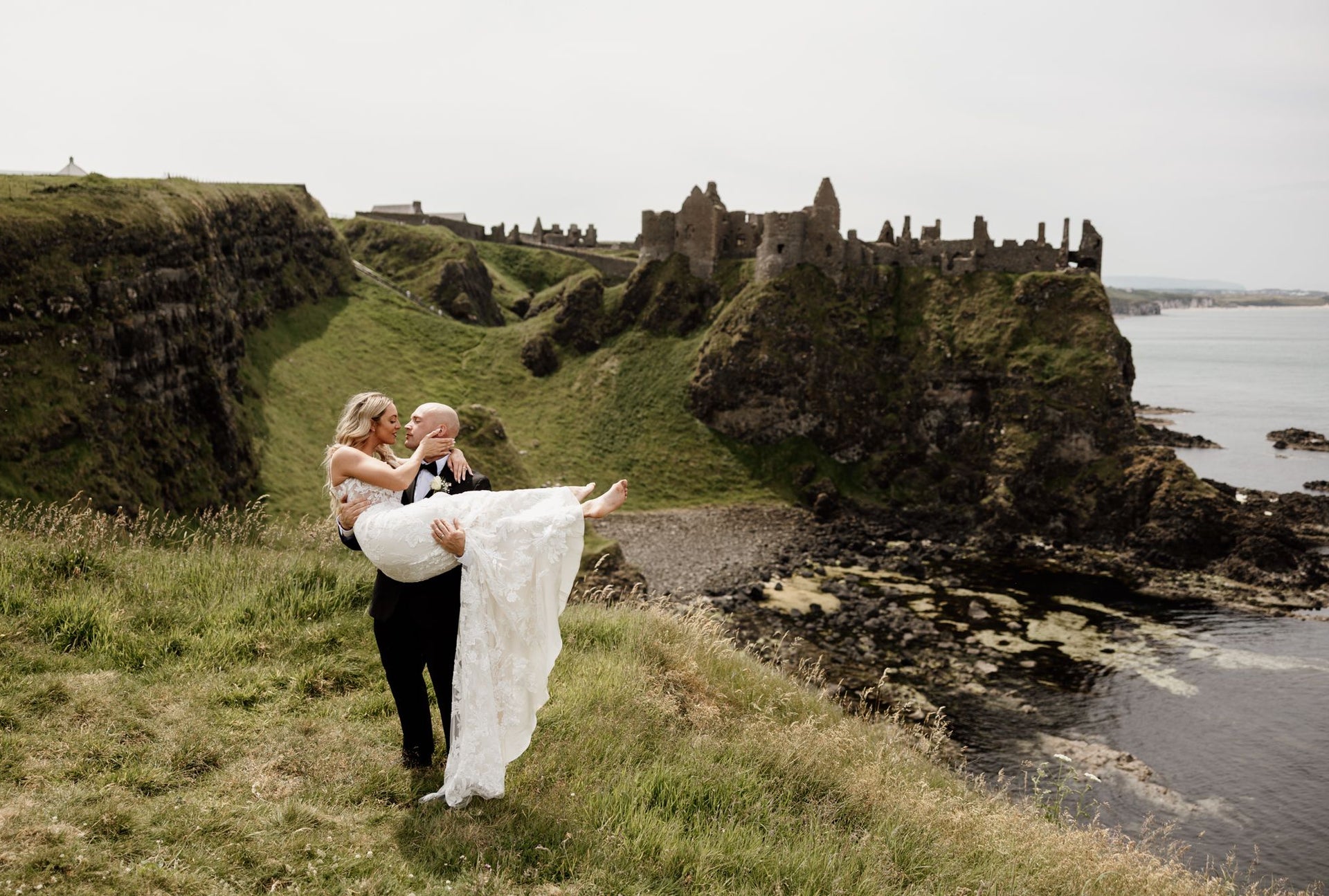 Groom holds bride in his arms with castle ruins and the sea in the background