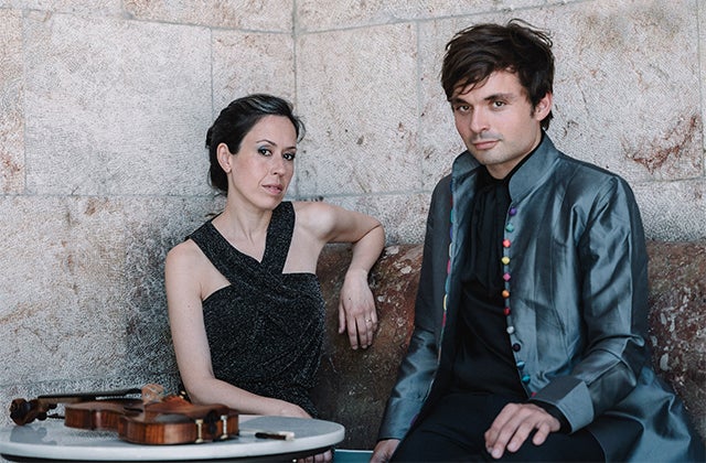 A woman in a black, sleeveless dress is seated beside a man in a dark grey shiny jacket with a violin lying on a small table in front of them against a tall grey stone background.