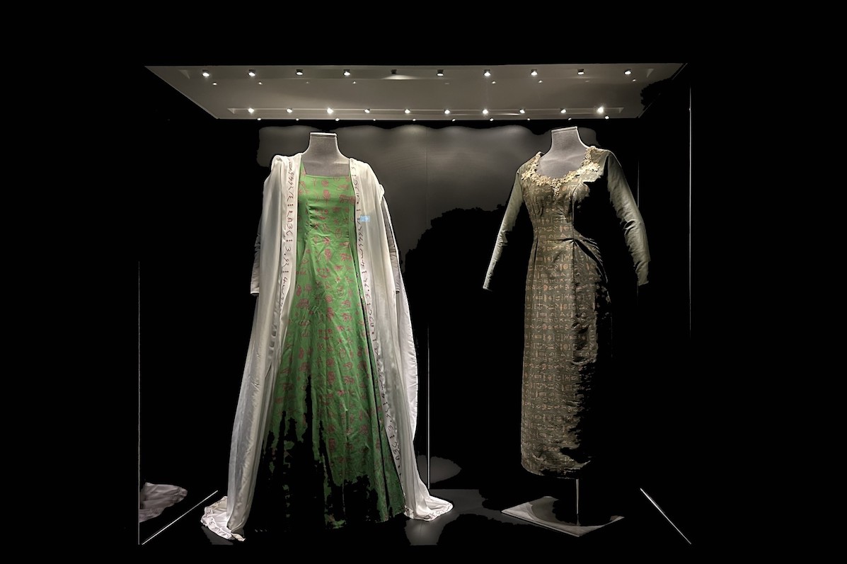 A dimly lit display cabinet with 2 full size displays of long dresses, against black background.
