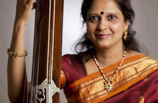 A smiling woman in a red and gold sari holding a tall, wooded, stringed musical instrument.