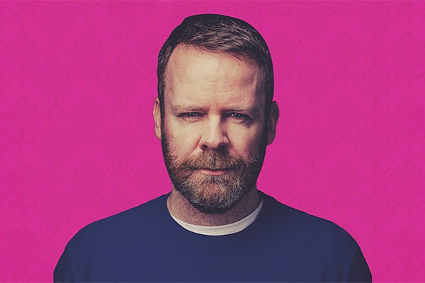 A man with a beard is looking slightly quizzical, in a dark blue jumper, against bright pink background.