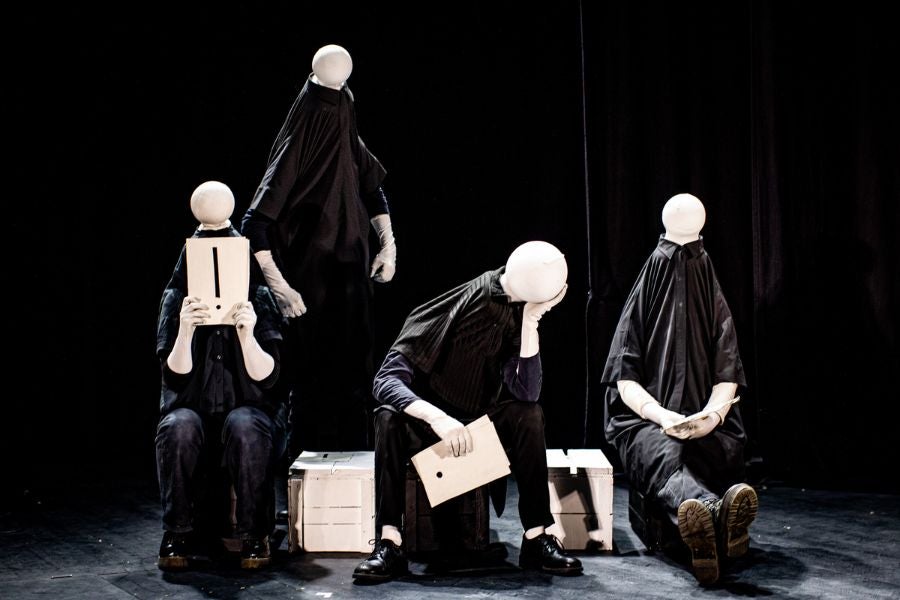4 plain figures on a stage dressed all in black with plain, small, round blank heads