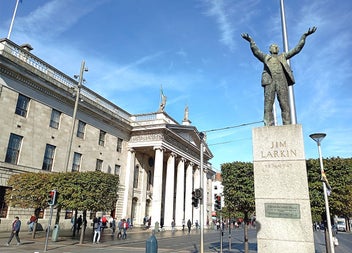 A statue on a plinth of a man standing with arms outstretched beside a busy street
