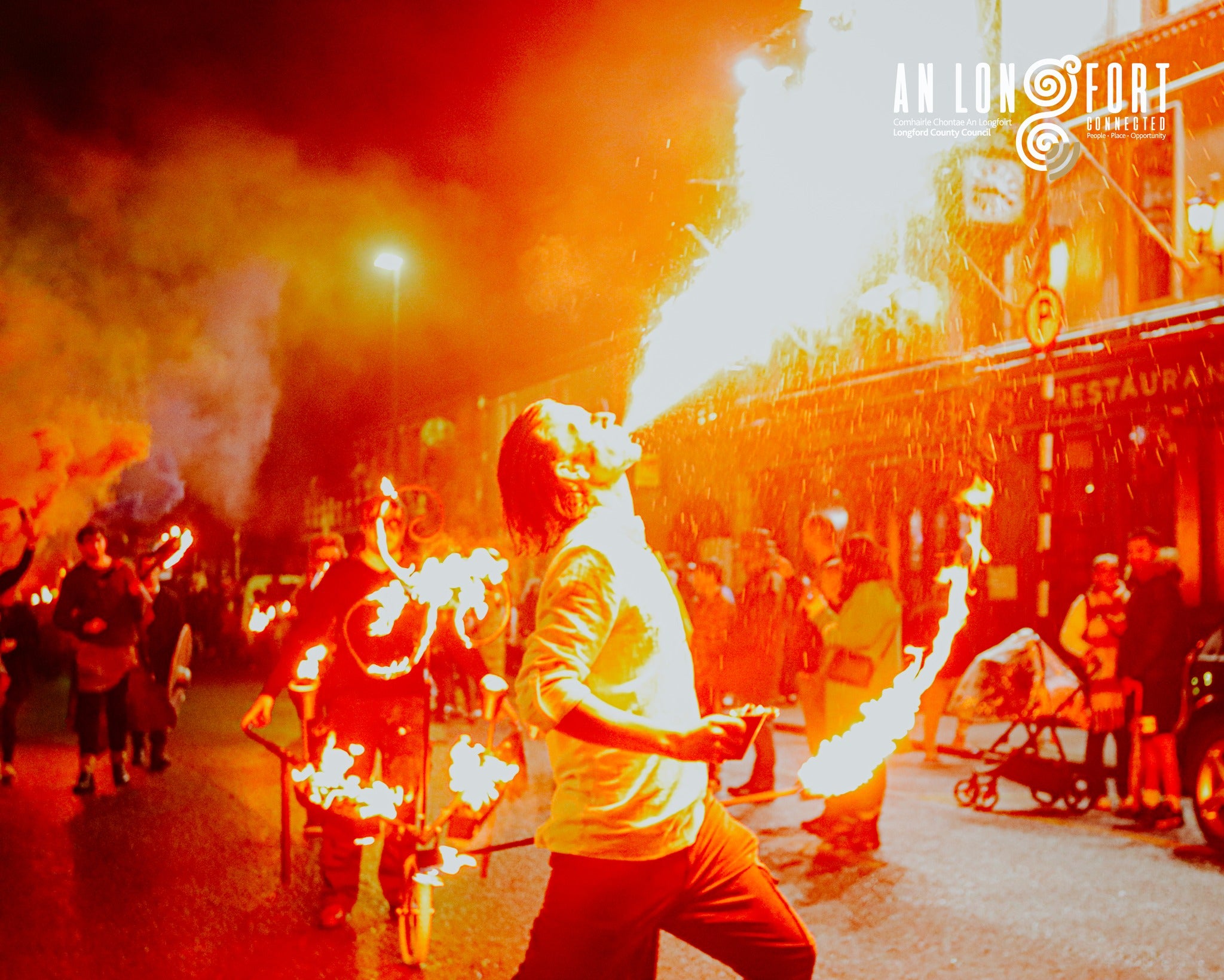 Orange coloured photo of person breathing fire with other lit objects in the background and people looking on.