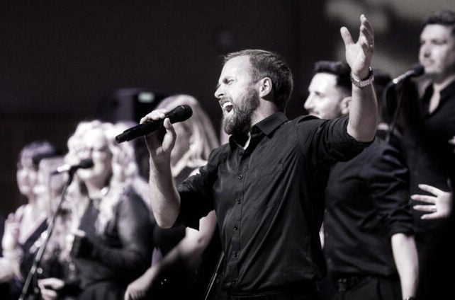 Black and white photo of part of large group singing, with central figure of man singing with emotion holding a mic.