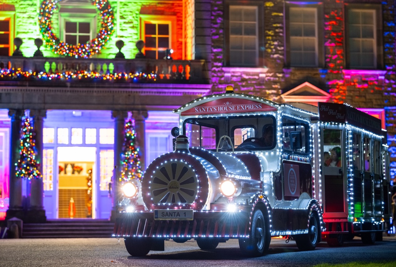 Santa's House Express at Palmerstown House. A train shaped vehicles pulling carriages at night, lit up in lots of tiny lights with illuminated large house behind.