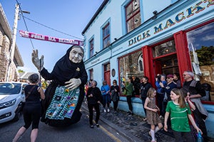 Large puppet of old woman in black as part of parade in a town street.