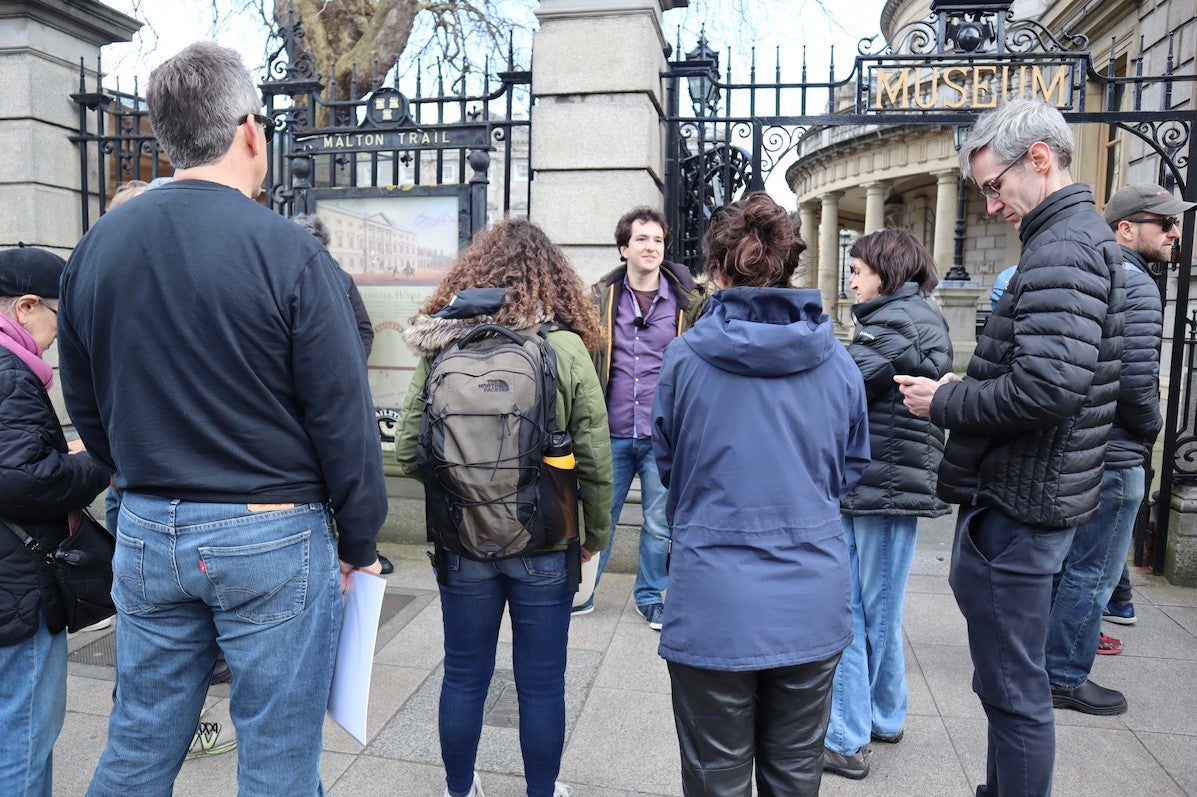 A small group of people are standing listening to a man standing outside tall railings and cream stone wall.