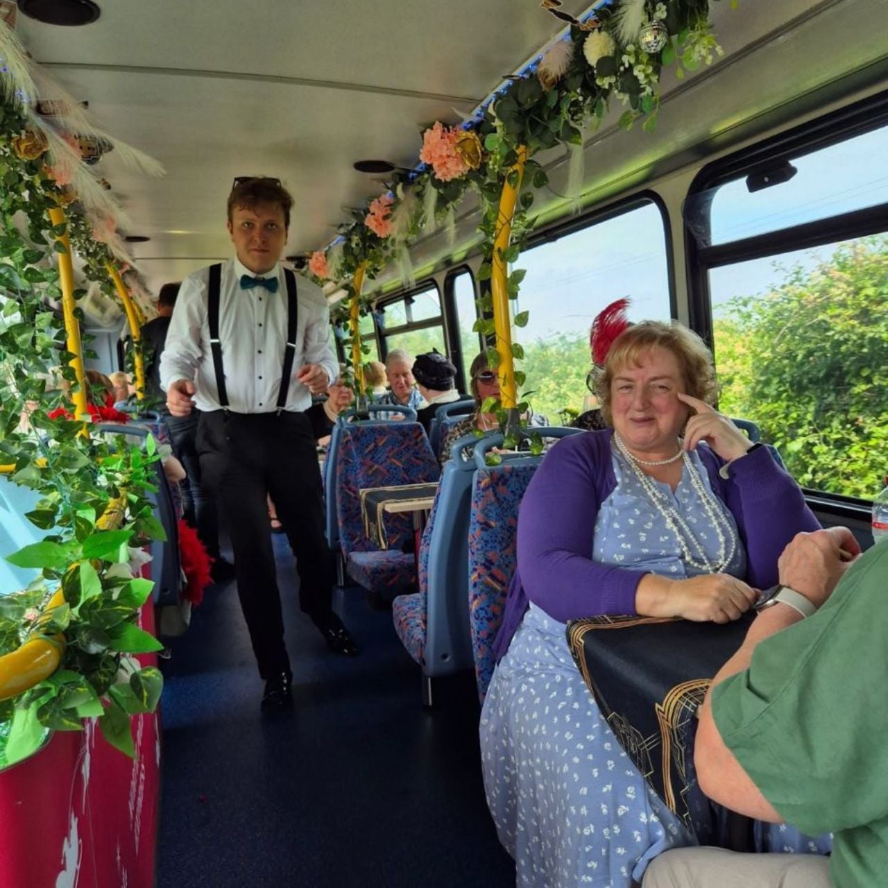 A man stands in the aisle of an Emerald Tea Tours bus with a woman smiling and sitting at a table by the window
