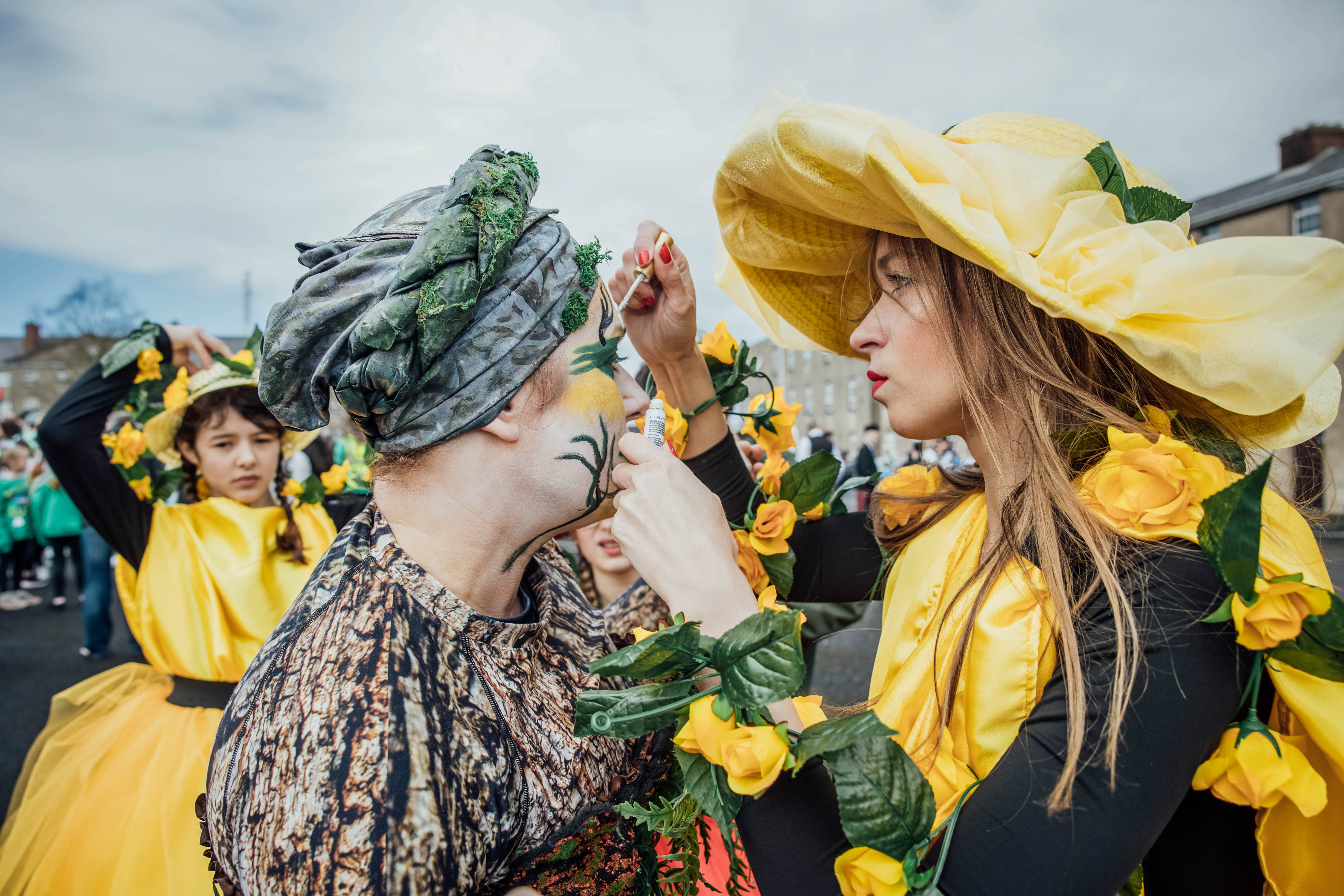 A person dressed in large yellow hat is applying colour to another person's face.