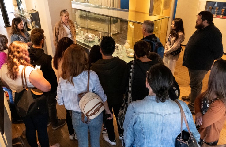 Seen from slightly above, a group of people are gathered around a glass display cabinet with a person standing beside it talking.