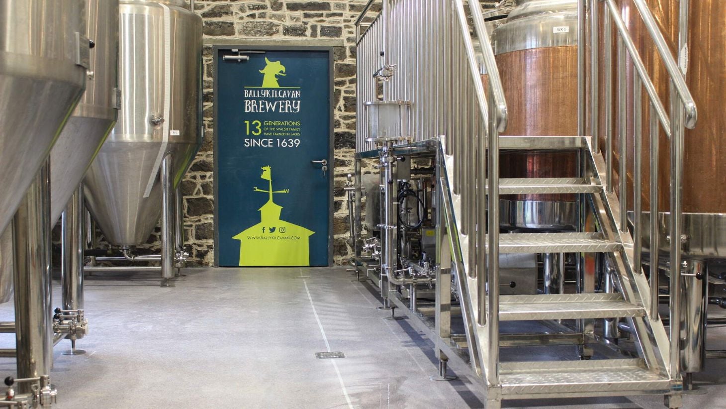 A picture of the inside of the Ballykilcavan Brewing Company with three large vats on the left and a blue door in the centre.