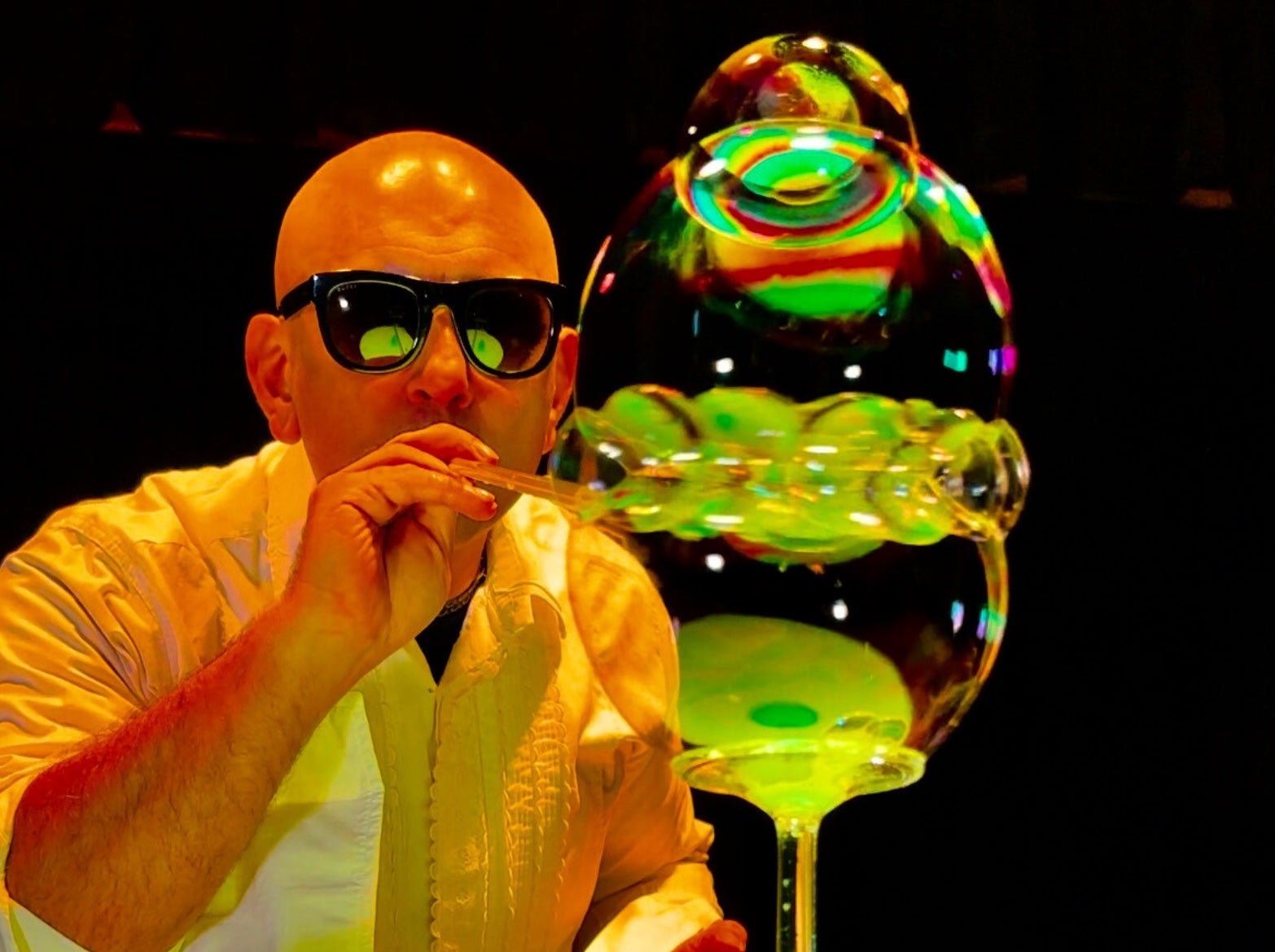 A man in dark glasses is blowing through a small tube with multiple large bubbles at the end