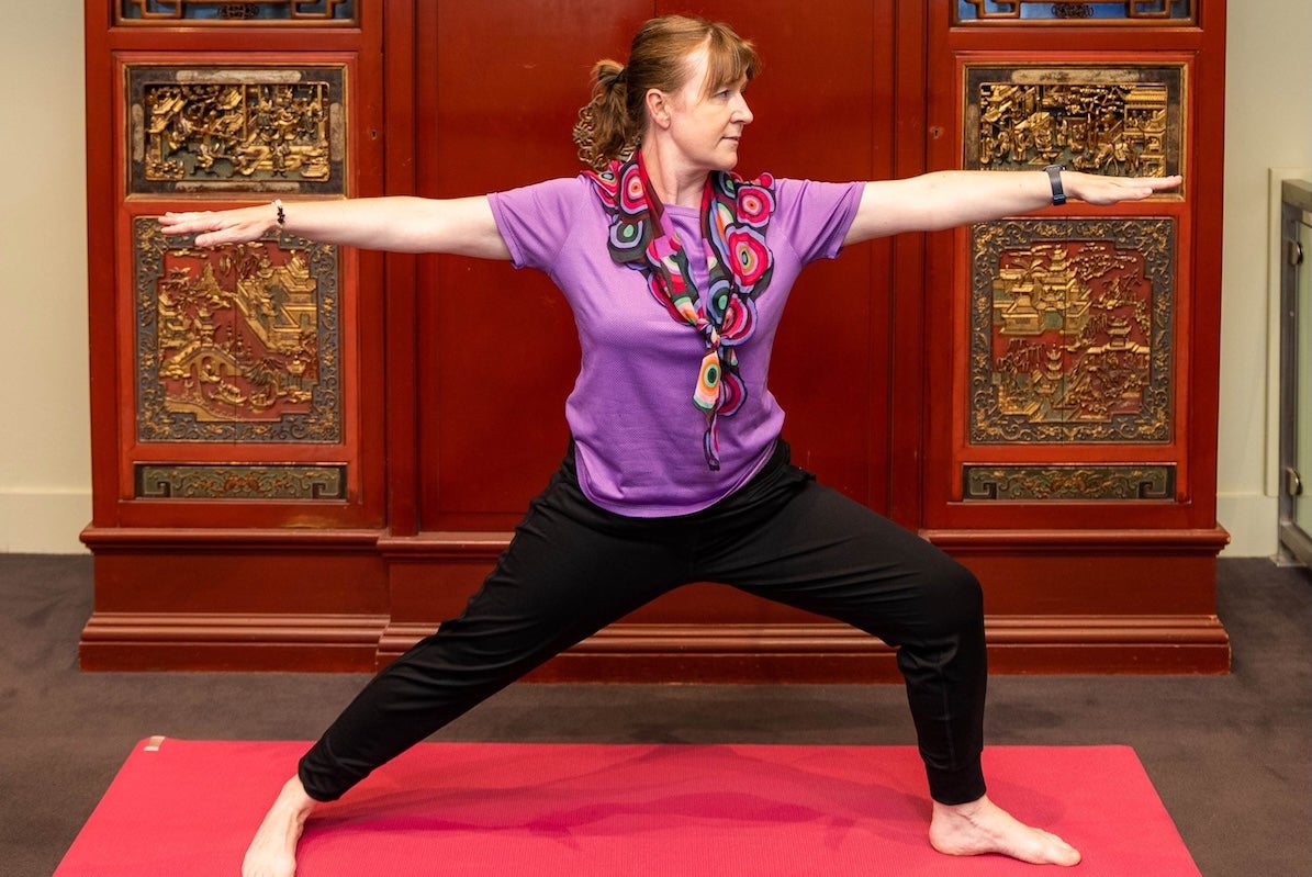 A woman with arms outstretched in a yoga pose with ornate dark red and gold cabinet behind her.