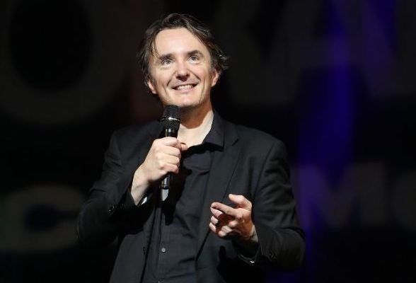 A smiling man holding a mic, gesturing with his left hand on a dark stage.