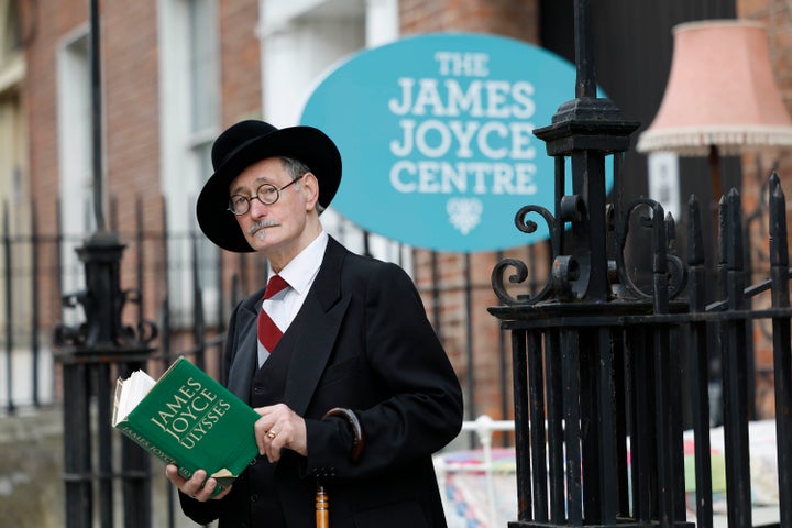 An elderly man in black suit and hat is holding open a green book, standing in front of black railings.