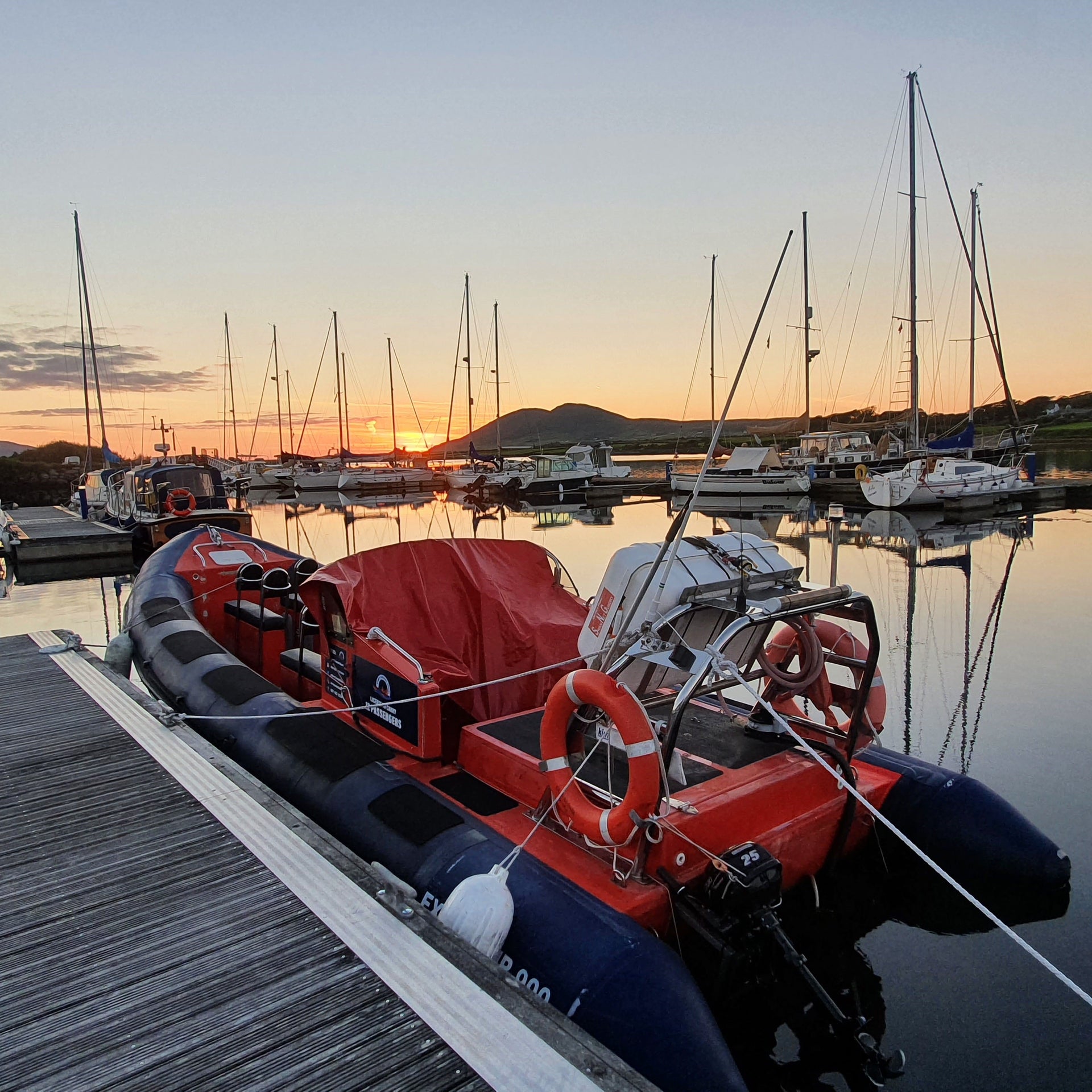 RIB boat docked in the water at a pier at sunset with sail boats and mountain landscape in the background