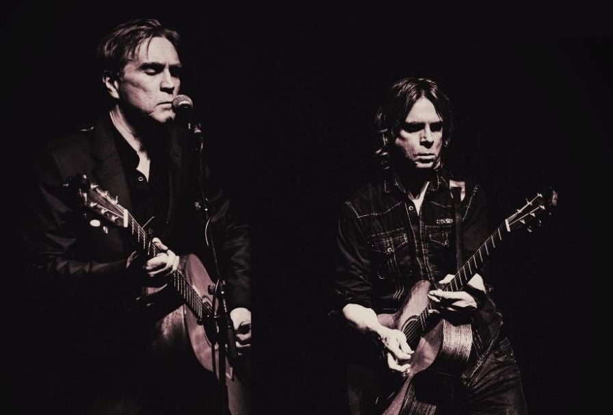 Darkly lit photo of 2 men, both playing guitar, one singing into mic on a stand, against black background.