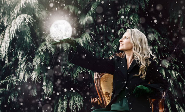 A woman in dark clothing is seated with a hand outstretched with a large, white glowing sphere on her flat palm against background of dark fur tree with snow.