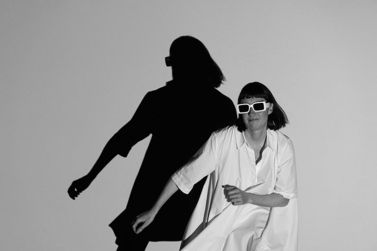 No Woman is an Island by Róisín Stack, a woman seated in dark sunglasses casting a large shadow of herself on plain wall behind.
