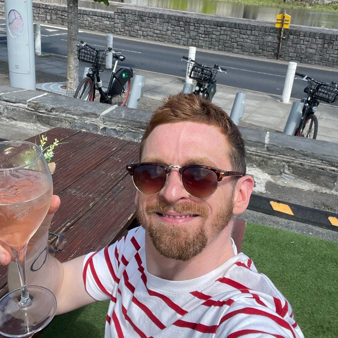 Man in a striped red and white shirt sitting at a bench table with bikes behind him, smiling as he holds up a drink to the camera.