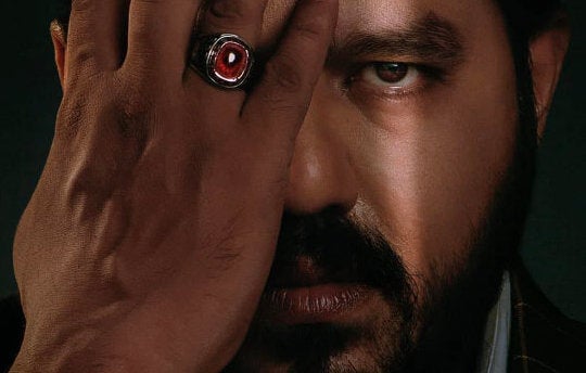 Close up view of a man's face with half covered by his hand wearing a red shiny ring.