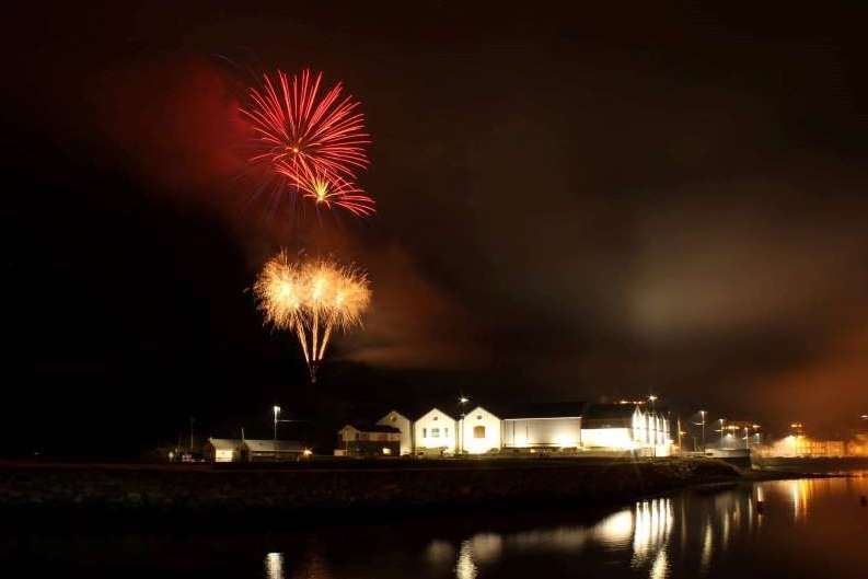 At night looking across water at a white building lit up with red and gold fireworks exploding in the dark sky above.