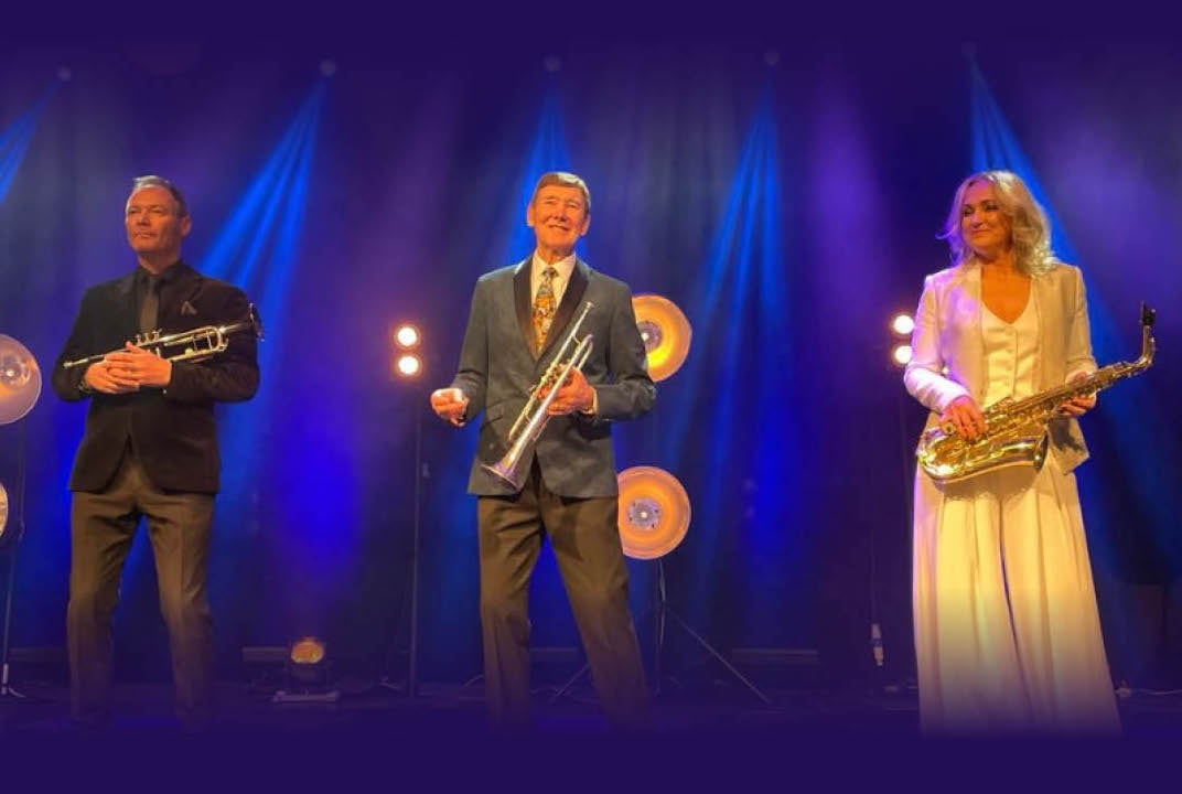 2 men holding brass musical instruments and a woman in a long yellow dress holding a saxophone are spread out in a line on a stage with blue lit background.