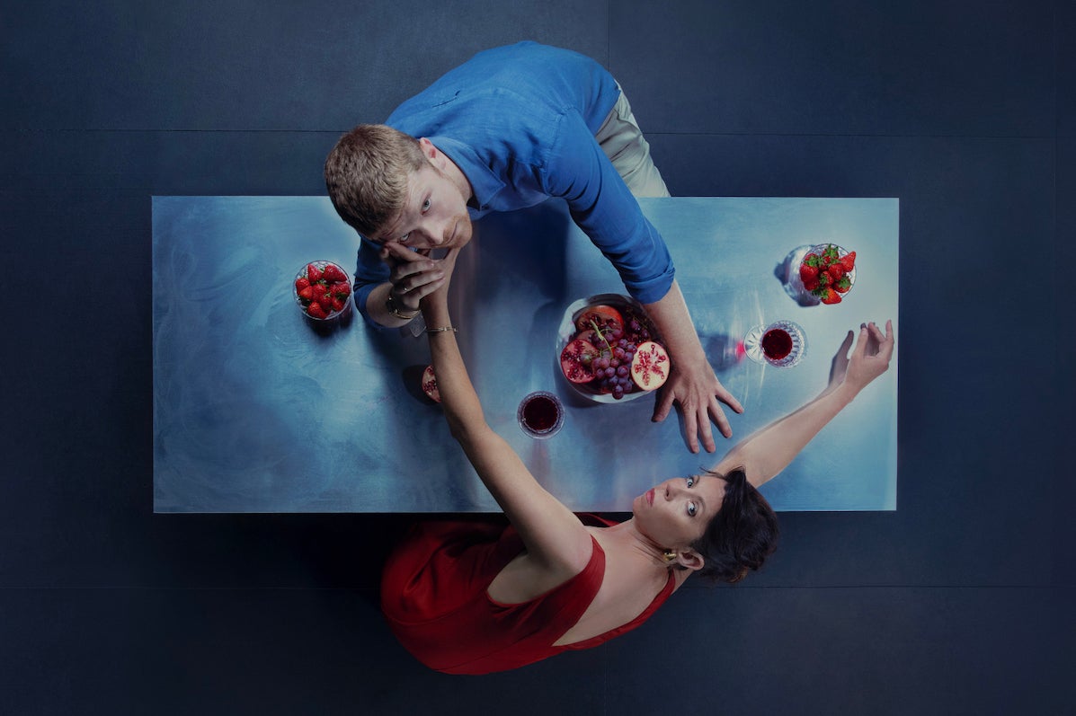 Viewed from above, a man and woman are reaching across a dinner table to each other, looking up at the viewer.